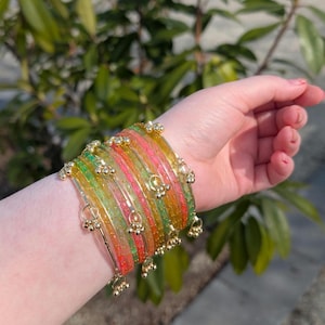 May include: A set of vibrant bangles in green, pink, orange, and yellow are displayed on a wrist. Each bangle is decorated with small gold-coloured charms. The background shows green leaves.