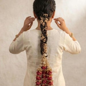 May include: A long, dark hair braid adorned with small gold-toned floral embellishments throughout the length. At the base of the braid, a large, decorative hair accessory features multiple deep red rose buds accented with gold-toned metallic details.