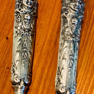 May include: Two antique silver-toned knife handles. Each handle features a detailed relief of a smiling face at the top, with ornate floral and grape vine designs. The handles are set against a warm, brown background.