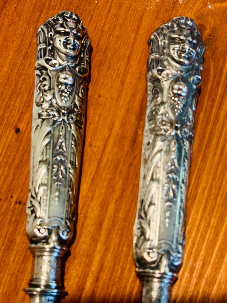 May include: Two antique silver-toned knife handles. Each handle features a detailed relief of a smiling face at the top, with ornate floral and grape vine designs. The handles are set against a warm, brown background.