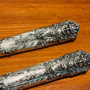 May include: Two decorative silver knife handles with detailed floral and fruit patterns. The handles have intricate textures and are set against a warm wooden background. The design includes grapes and leaves, typical of antique cutlery.