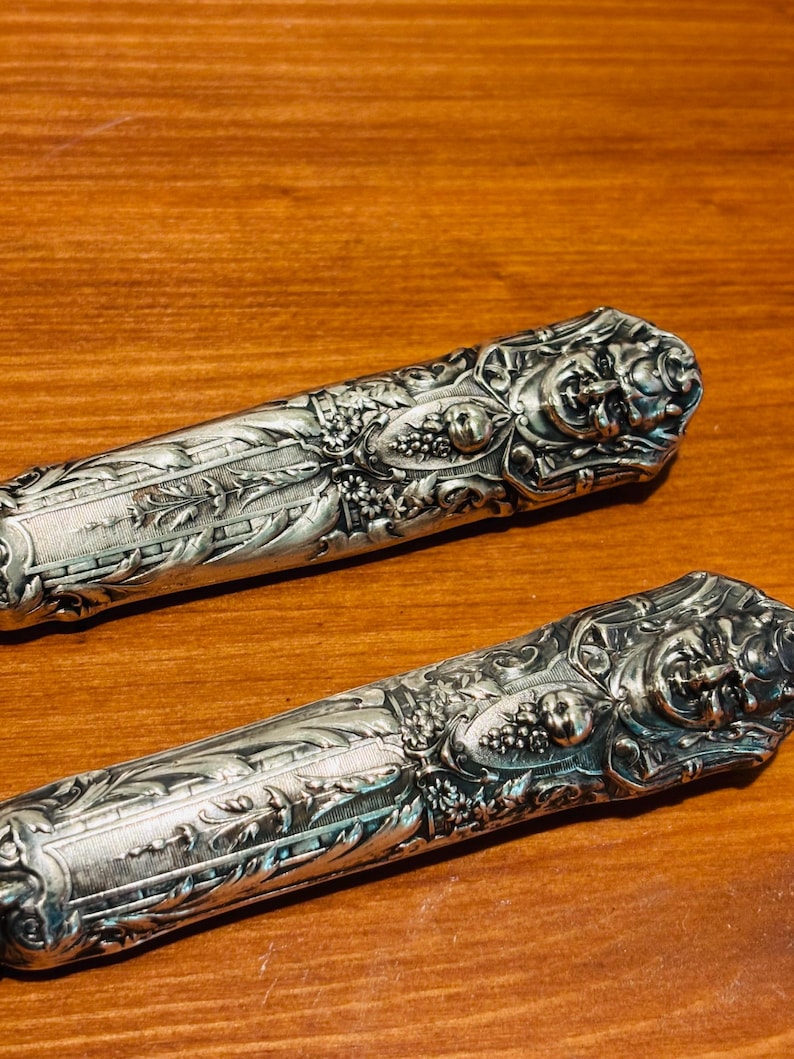 May include: Two decorative silver knife handles with detailed floral and fruit patterns. The handles have intricate textures and are set against a warm wooden background. The design includes grapes and leaves, typical of antique cutlery.