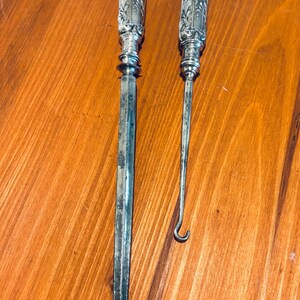 May include: Two antique silver tools with ornate handles. One is a letter opener, the other a hook-shaped tool. Both have detailed floral and face designs on the handles. The tools are displayed on a wooden surface.