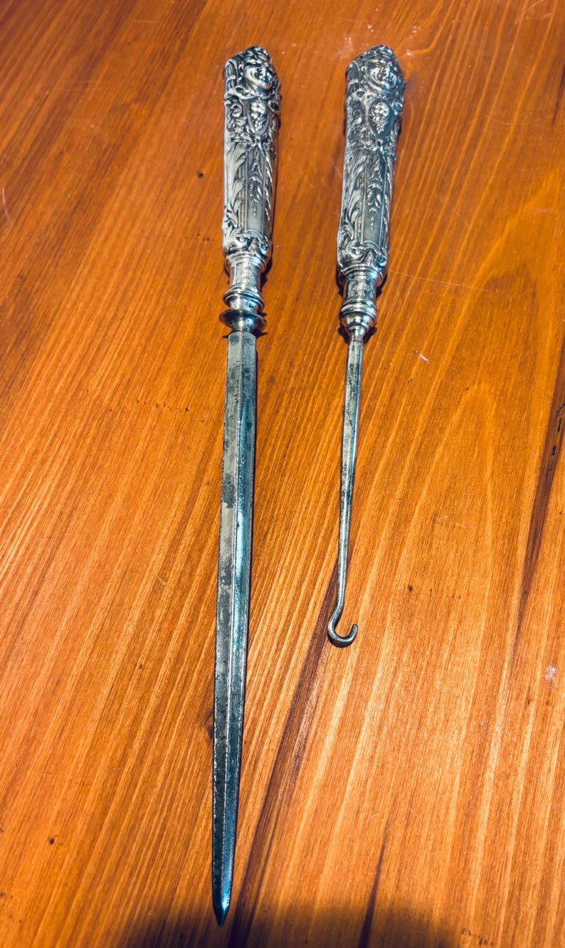 May include: Two antique silver tools with ornate handles. One is a letter opener, the other a hook-shaped tool. Both have detailed floral and face designs on the handles. The tools are displayed on a wooden surface.