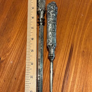 May include: Two antique silver tools with ornate handles. The longer tool is a letter opener, approximately 30 cm long. The shorter tool has a curved end, possibly a button hook. Both tools are on a wooden surface.