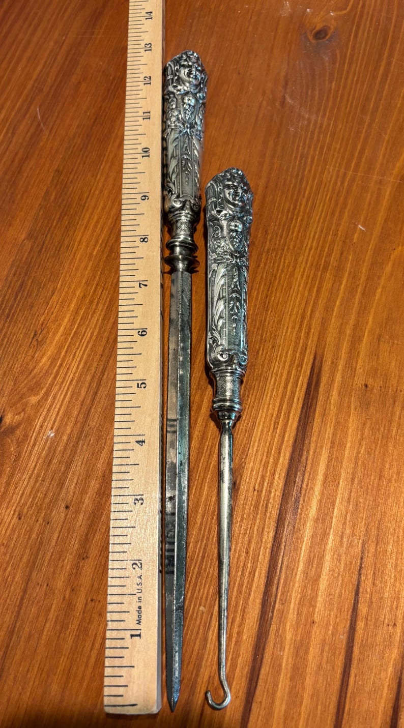 May include: Two antique silver tools with ornate handles. The longer tool is a letter opener, approximately 30 cm long. The shorter tool has a curved end, possibly a button hook. Both tools are on a wooden surface.