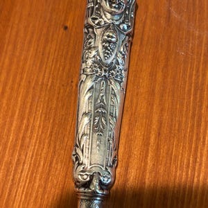 May include: An ornate silver knife handle with detailed relief carvings. The handle features a smiling face at the top, a shield with grapes, and intricate floral and scrollwork designs. The handle is approximately 15 cm long.