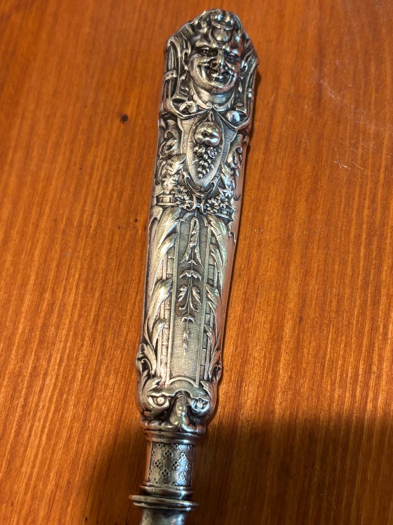 May include: An ornate silver knife handle with detailed relief carvings. The handle features a smiling face at the top, a shield with grapes, and intricate floral and scrollwork designs. The handle is approximately 15 cm long.
