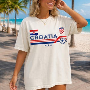 Puede incluir: Una camiseta de manga corta color hueso con el texto CROATIA en azul, acompa&ntilde;ado por la bandera de Croacia, el escudo nacional, un gr&aacute;fico de un bal&oacute;n de f&uacute;tbol y tres estrellas azules, todo dispuesto con rayas horizontales rojas y azules.