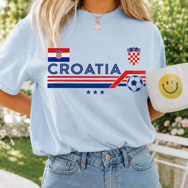 Puede incluir: Una camiseta azul claro con el texto CROATIA en letras azules, una peque&ntilde;a bandera croata, el escudo de armas de Croacia, un gr&aacute;fico de un bal&oacute;n de f&uacute;tbol y tres estrellas azules. El dise&ntilde;o incluye rayas horizontales rojas y azules en el pecho.