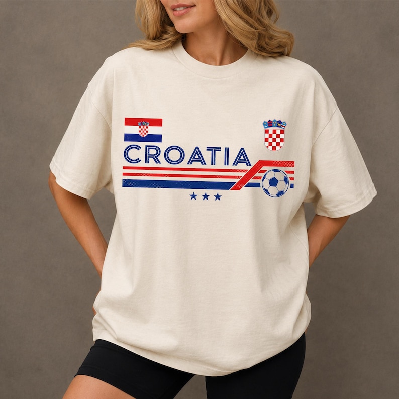 Puede incluir: Una camiseta de manga corta color hueso con el texto CROATIA en azul, una bandera de Croacia, un escudo ajedrezado rojo y blanco, rayas horizontales rojas y azules, un gr&aacute;fico de un bal&oacute;n de f&uacute;tbol y tres estrellas azules bajo el texto.
