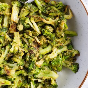Op de afbeelding: Een kom met geroosterde broccoliroosjes, lichtgroen van kleur en met lichtbruine randen. De broccoli is gemengd met een saus en wordt geserveerd in een gespikkelde witte kom met een zilveren lepel.