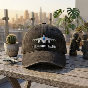 May include: A charcoal gray distressed baseball cap featuring a white graphic of an F-16 Fighting Falcon aircraft centered on the front panel, with the text F-16 FIGHTING FALCON printed in white capital letters directly below the airplane graphic.