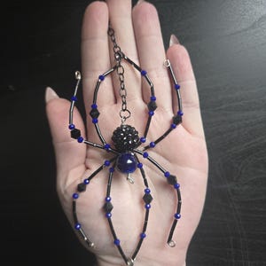 May include: A beaded spider pendant with a black body and legs, accented with blue and black beads. The spider has a chain for hanging. The spider's body is a dark blue color. The pendant is held in a hand.
