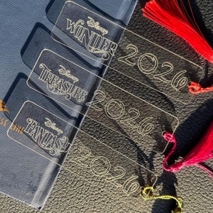 May include: Clear acrylic bookmarks with red tassels. Each bookmark features the Disney Cruise Line logo and the words "WONDER", "TREASURE", and "FANTASY" along with the year "2026". The bookmarks are on a dark blue surface.