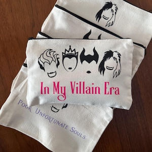 May include: Off-white canvas cosmetic bags with black trim and zippers. The bags feature black silhouettes of Disney villains and the pink text "In My Villain Era." One bag has the text "Poor Unfortunate Souls."