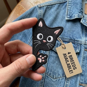 May include: Black cat-shaped embroidered patch with pink accents on the ears and paw. The patch has white eyes and whiskers. A tag attached to the patch reads "4 BREEDS AVAILABLE!" The patch is held against a denim jacket.