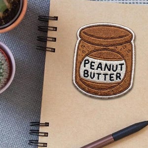 May include: A brown embroidered patch in the shape of a peanut butter jar with the words "PEANUT BUTTER" in white. The patch is on a spiral-bound notebook. A pen and two potted cacti are also visible.