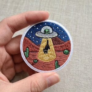 May include: Embroidered patch featuring a black cat being abducted by a UFO. The design includes a blue starry sky, red desert landscape, green cacti, and a yellow beam of light. The patch is round with a white border.