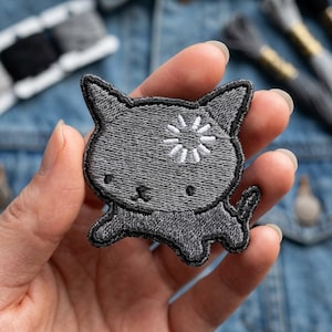 May include: Embroidered gray cat patch with a white floral design on its forehead. The patch has a black outline and is in the shape of a cartoon cat. The patch is held in a person's hand.