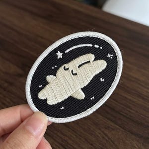 May include: Embroidered patch featuring a cartoon space bunny design. The patch has a black oval background with white stars and a curved line. The bunny is light beige with a smiling face. The patch is outlined with white stitching.