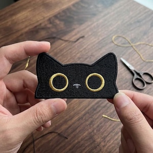 May include: Black cat patch with gold embroidered eyes and details. The patch is held between two hands, with scissors and thread in the background. The cat patch is a cute, whimsical design.