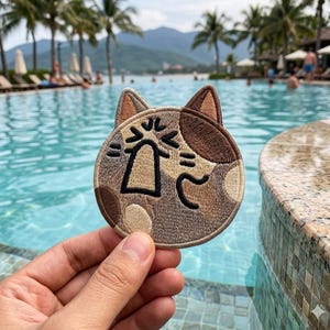 May include: Embroidered patch featuring a cat face with a surprised expression. The patch is round, with a tan and brown color scheme, and black embroidered details. The cat's face has large eyes and a wide open mouth. The patch is held in front of a pool.