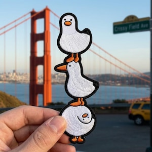 May include: Embroidered patch featuring three stacked cartoon ducks. The ducks are white with black outlines, orange feet and beaks. The background includes the Golden Gate Bridge and a street sign that reads "Crissy Field Ave."