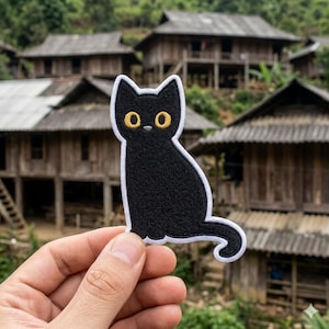 May include: A black cat patch with yellow eyes and a white border. The cat is sitting with its tail curled. The patch is held in front of a blurred background of wooden buildings and green foliage.