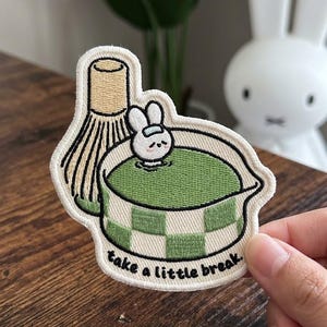 May include: Embroidered patch featuring a bunny relaxing in a matcha bath, with a matcha whisk. The patch has a green and white checkered pattern and the text "take a little break."