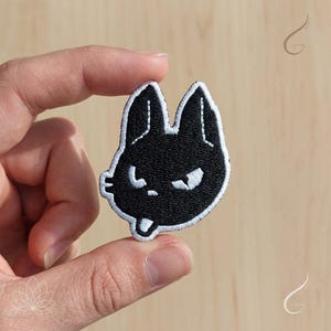 May include: Black and white embroidered patch featuring a stylized rabbit face with an angry expression. The patch has a black background with white stitching outlining the rabbit's features, including its ears, eyes, and mouth. The patch is held between two fingers.