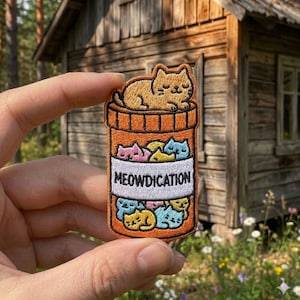 May include: Embroidered patch featuring a pill bottle design with the word "MEOWDICATION" on a white label. The bottle is filled with colorful cat faces, and a larger cat rests on top. The patch is orange, with brown and various other colors.