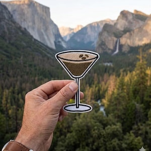 May include: Embroidered patch featuring a coffee martini design with a brown drink and coffee beans. The patch has a white border and is held in front of a scenic mountain landscape with green trees and a waterfall.