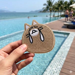 May include: A brown embroidered patch in the shape of a cat's face with a sad expression. The cat's face has white accents and black embroidered details. The patch is held in a person's hand, with a pool and ocean in the background.
