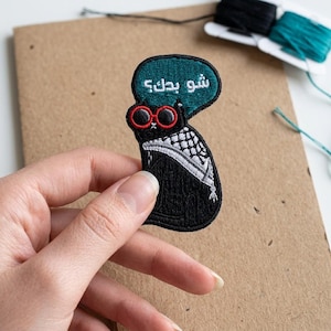 May include: Embroidered black cat patch with red sunglasses and a teal speech bubble. The speech bubble contains white text. The cat wears a black and white scarf. Sewing thread spools are in the background.