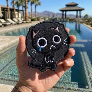 May include: Black embroidered patch featuring a cartoon cat with large white eyes, a blue teardrop, and a sad expression. The cat has pink accents on its ears and whiskers. The patch is round and has a black border.