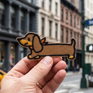 May include: A brown dachshund patch with black and yellow accents. The embroidered patch features a smiling dog with small orange wings. The patch is held in front of a city street scene.