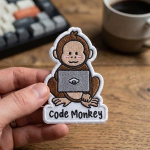 Puede incluir: Parche bordado con un mono de dibujos animados con un portátil, con la etiqueta "Code Monkey". El mono es marrón con un portátil gris y un borde blanco. El parche se sostiene en una mano sobre una superficie de madera.