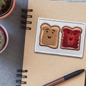 May include: Embroidered patch featuring two slices of toast with smiling faces. One slice is peanut butter, the other is jelly. The patch is rectangular with a white border. The patch is on a spiral notebook with a pen.