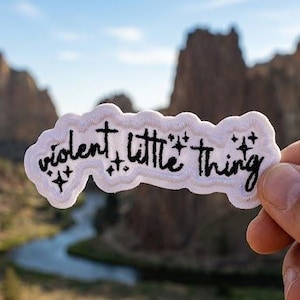 May include: A white sticker with the words "violent, little thing" in black script, accented with small star graphics. The sticker is held in front of a scenic outdoor background featuring mountains and a river.