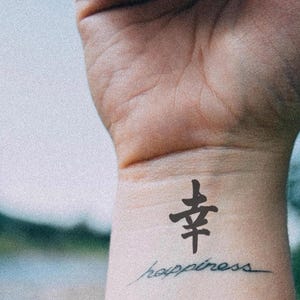 May include: A wrist with a black tattoo of a Chinese character and the word "happiness" in cursive script. The skin tone is light, and the background is blurred with shades of blue and green.