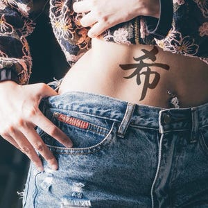 May include: A person wearing a floral patterned jacket and denim shorts. A black tattoo of a Japanese character is visible on the abdomen. The shorts have a frayed hem and visible pockets.