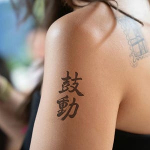 Puede incluir: Primer plano de un hombro con un tatuaje negro de dos caracteres chinos. El tatuaje está en la parte superior del brazo. Un pequeño tatuaje descolorido es visible en el hombro. La persona lleva una camiseta negra.