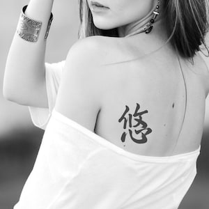 Puede incluir: Imagen en blanco y negro de la espalda y el hombro de una persona. Un tatuaje negro de un carácter chino es visible en el hombro. La persona lleva un top blanco con hombros descubiertos y una pulsera de puño plateada.