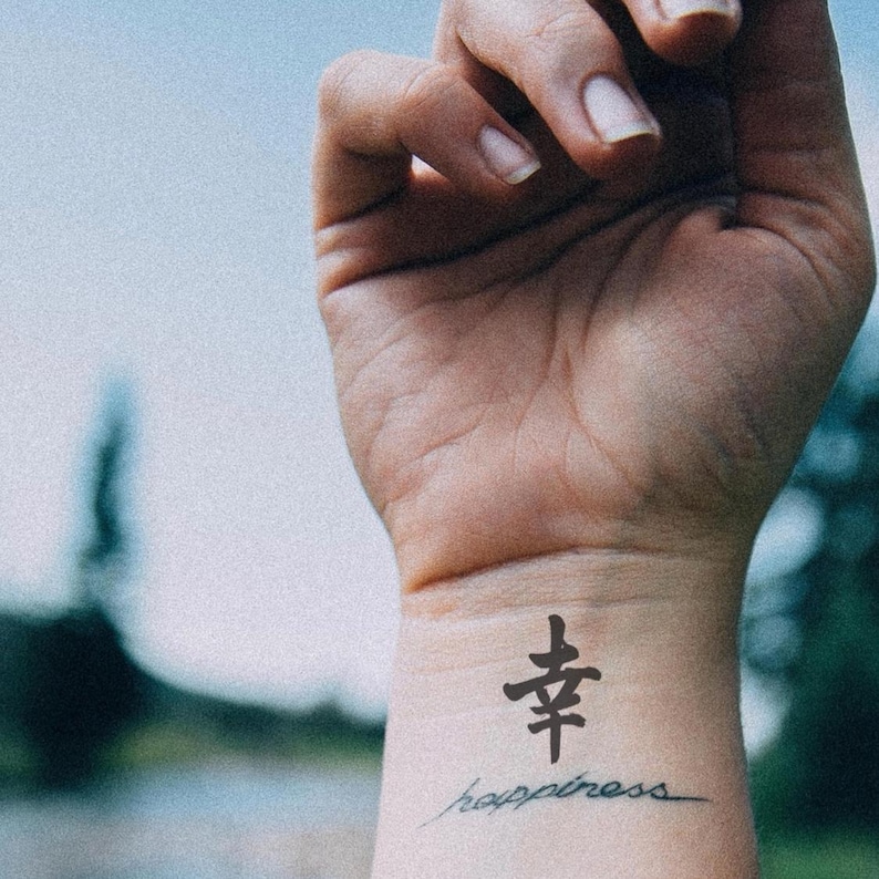 May include: A wrist with a black tattoo of a Chinese character and the word "happiness" in cursive script. The skin tone is light, and the background is blurred with shades of blue and green.