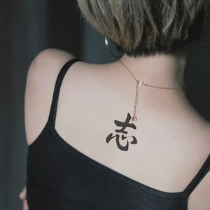 以下が含まれることがあります： 細いストラップが付いた黒いタンクトップ。ペンダント付きのゴールドネックレスが首の後ろにぶら下がっています。背中には漢字の黒いタトゥーが見えます。