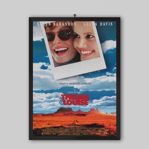 Puede incluir: Póster enmarcado de la película "Thelma & Louise" con Susan Sarandon y Geena Davis. El póster tiene una foto de las actrices, el título y un paisaje desértico. El póster está en un marco negro.
