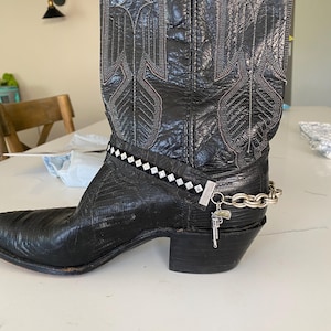 May include: Black leather cowboy boot with decorative stitching and a silver chain with a gun charm. The boot has a pointed toe and a stacked heel. A decorative band with white and black accents is around the ankle.