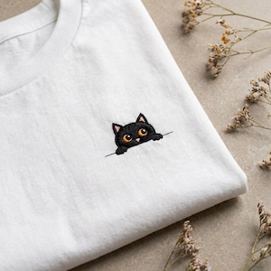 Può includere: T-shirt bianco con un ricamo di un gatto nero che sbuca da una linea. Il gatto ha occhi arancioni e orecchie rosa. Il disegno è sul lato sinistro della maglietta. Fiori secchi sullo sfondo.