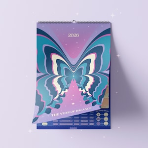 May include: A 2026 wall calendar with a butterfly design in shades of blue, purple, and pink. The calendar is titled "THE YEAR OF BALANCE" and features a spiral binding at the top. The design is symmetrical and artistic.